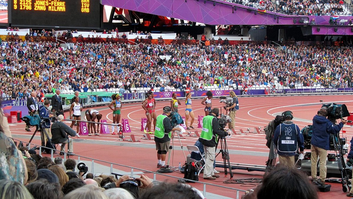 Sprinters in colorful kits stand beside numbered starting blocks on a red track in a packed Olympic stadium, surrounded by camera operators