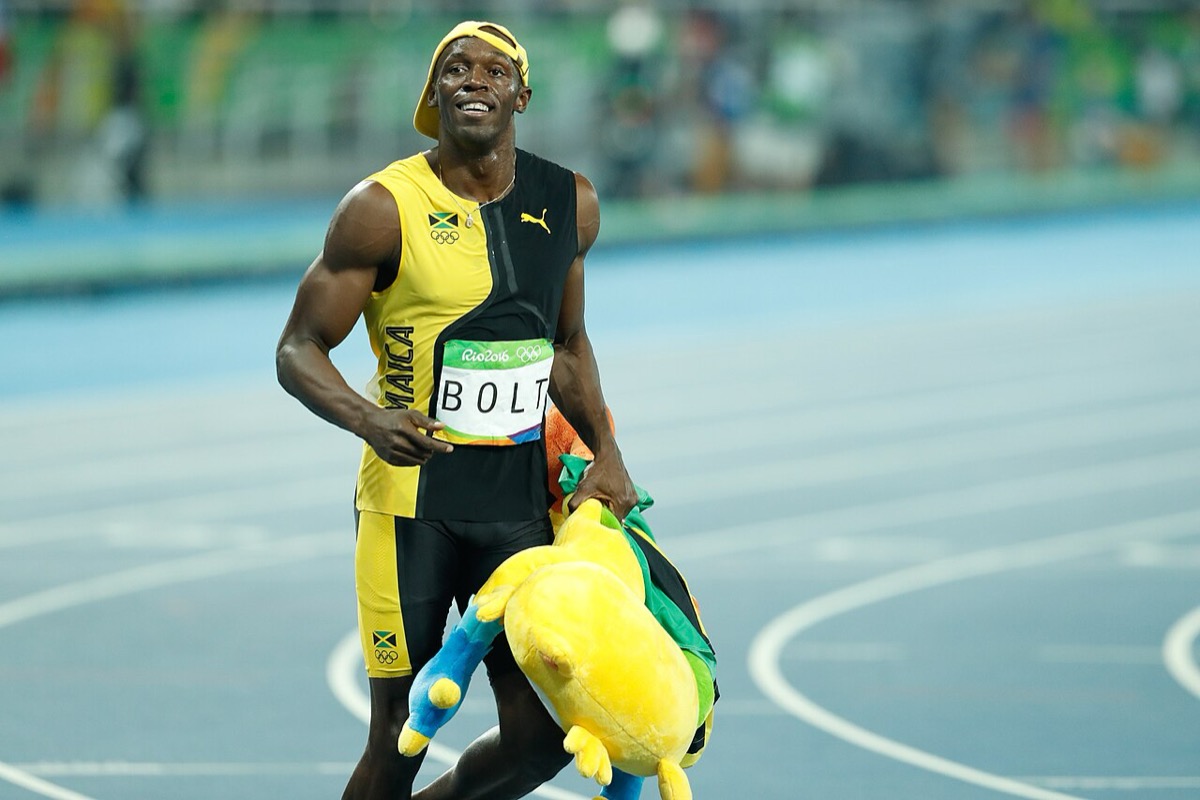 Usain Bolt in a yellow Jamaica kit walks along a blue track carrying a large yellow stuffed toy, smiling