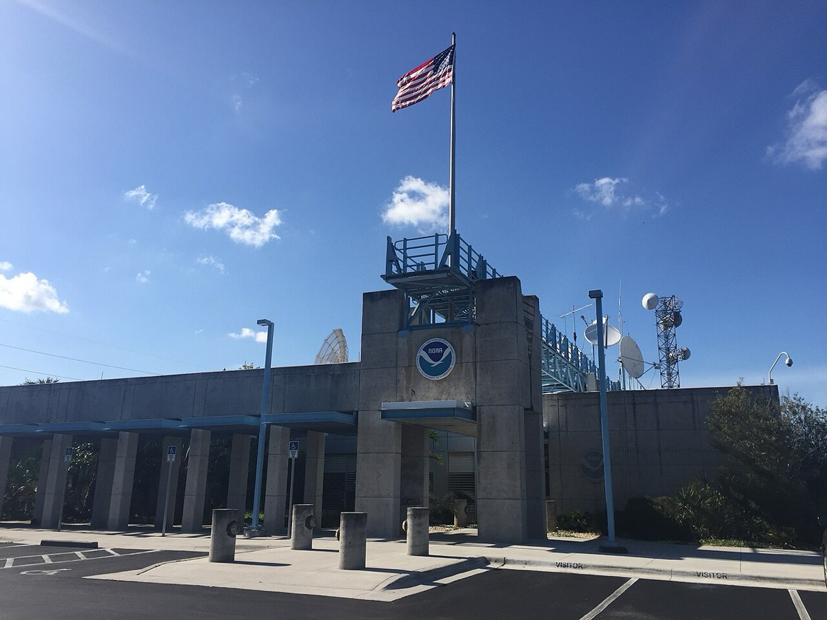 Concrete entrance of the National Hurricane Center with a flagpole, a NOAA emblem and satellite dishes on the roof under a blue sky